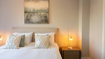 A neatly made bed with white linens and decorative pillows, a bedside table with a lamp, and a painting on the wall.