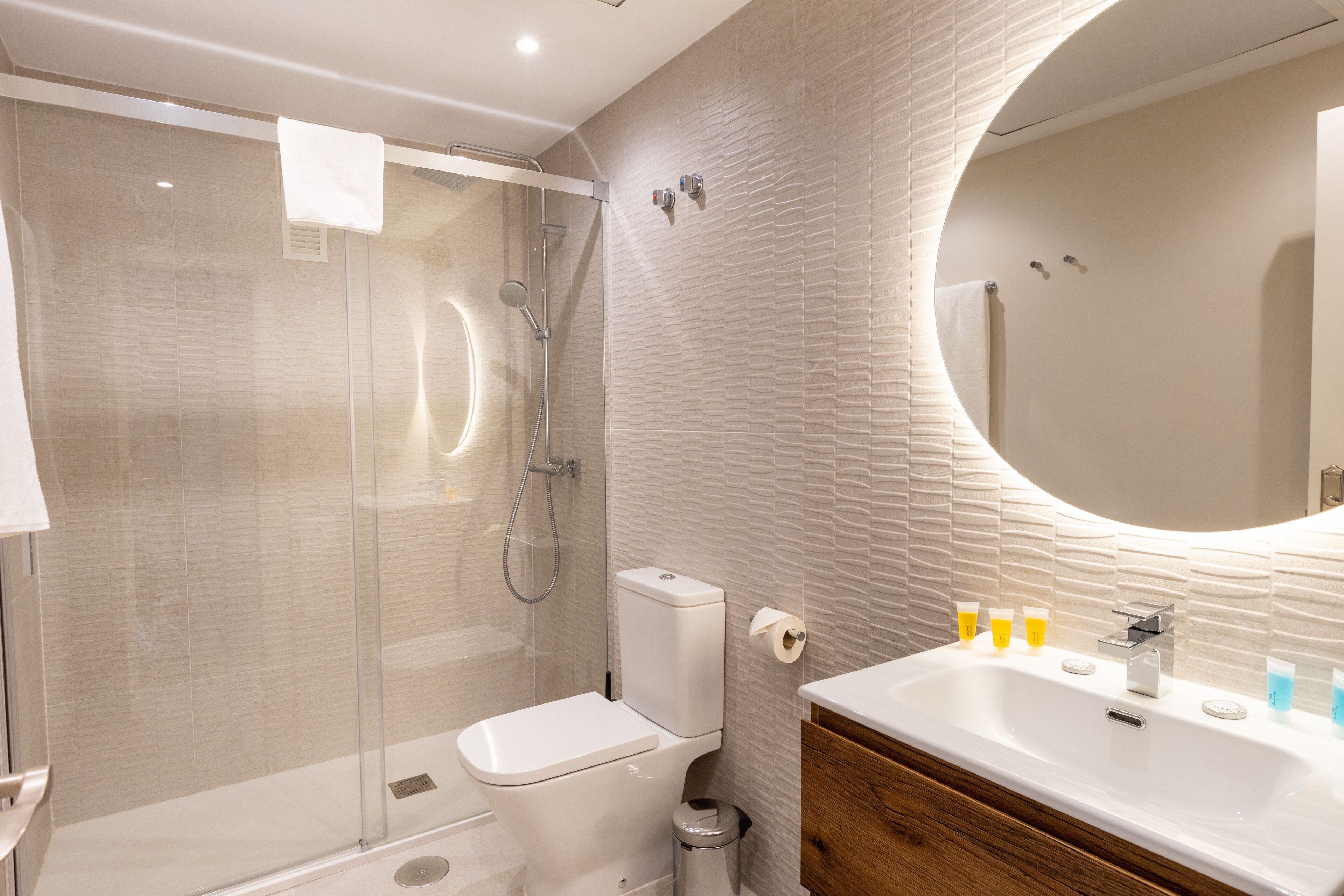 A modern bathroom with a glass-enclosed shower, a toilet, a sink with toiletries, and a mirror.