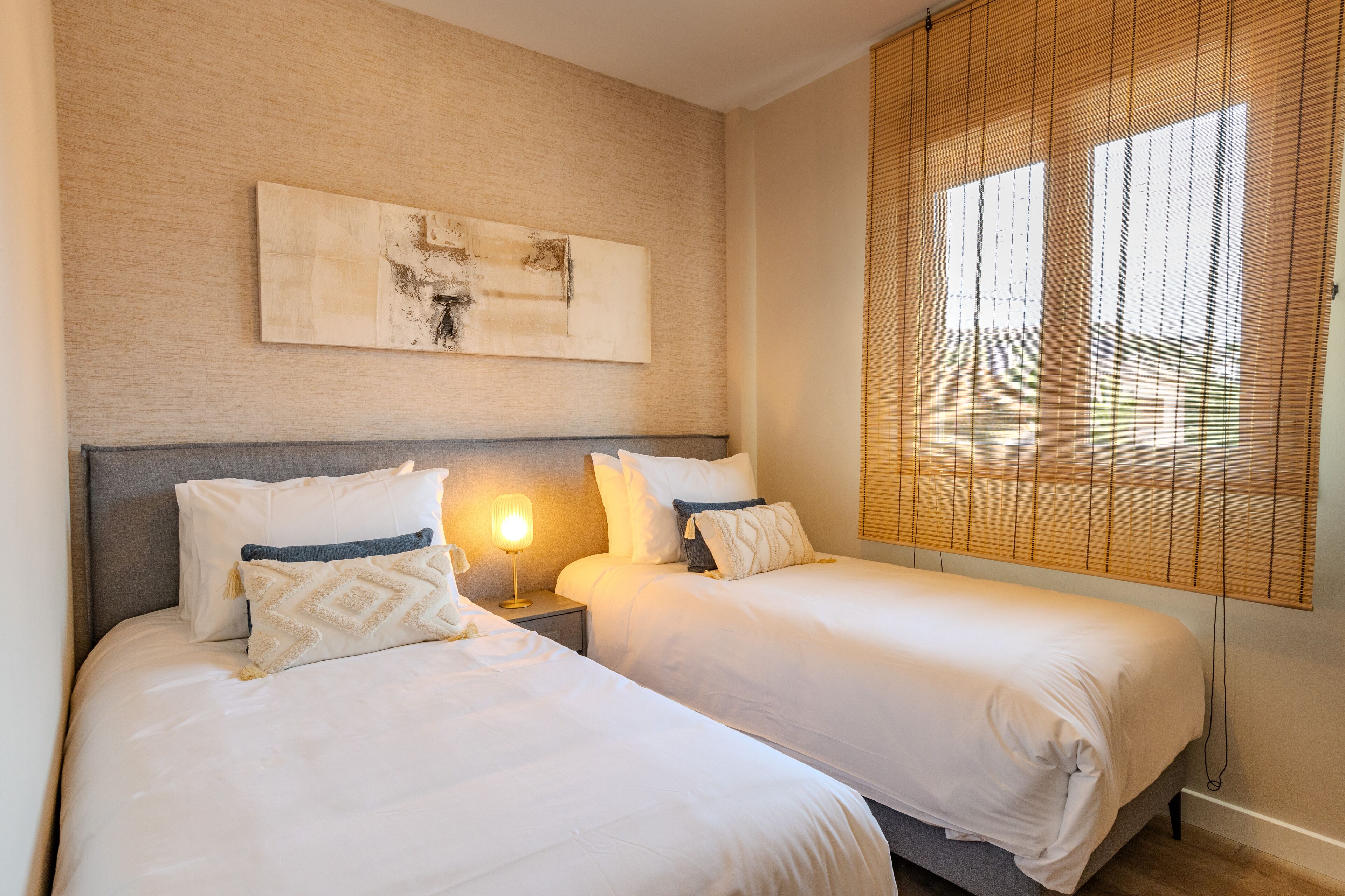 Une chambre d’hôtel avec deux lits, une table de chevet avec une lampe, une œuvre d’art accrochée au mur et une fenêtre avec des stores.