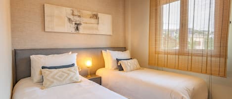A hotel room with two beds, a nightstand with a lamp, a wall-mounted artwork, and a window with blinds.