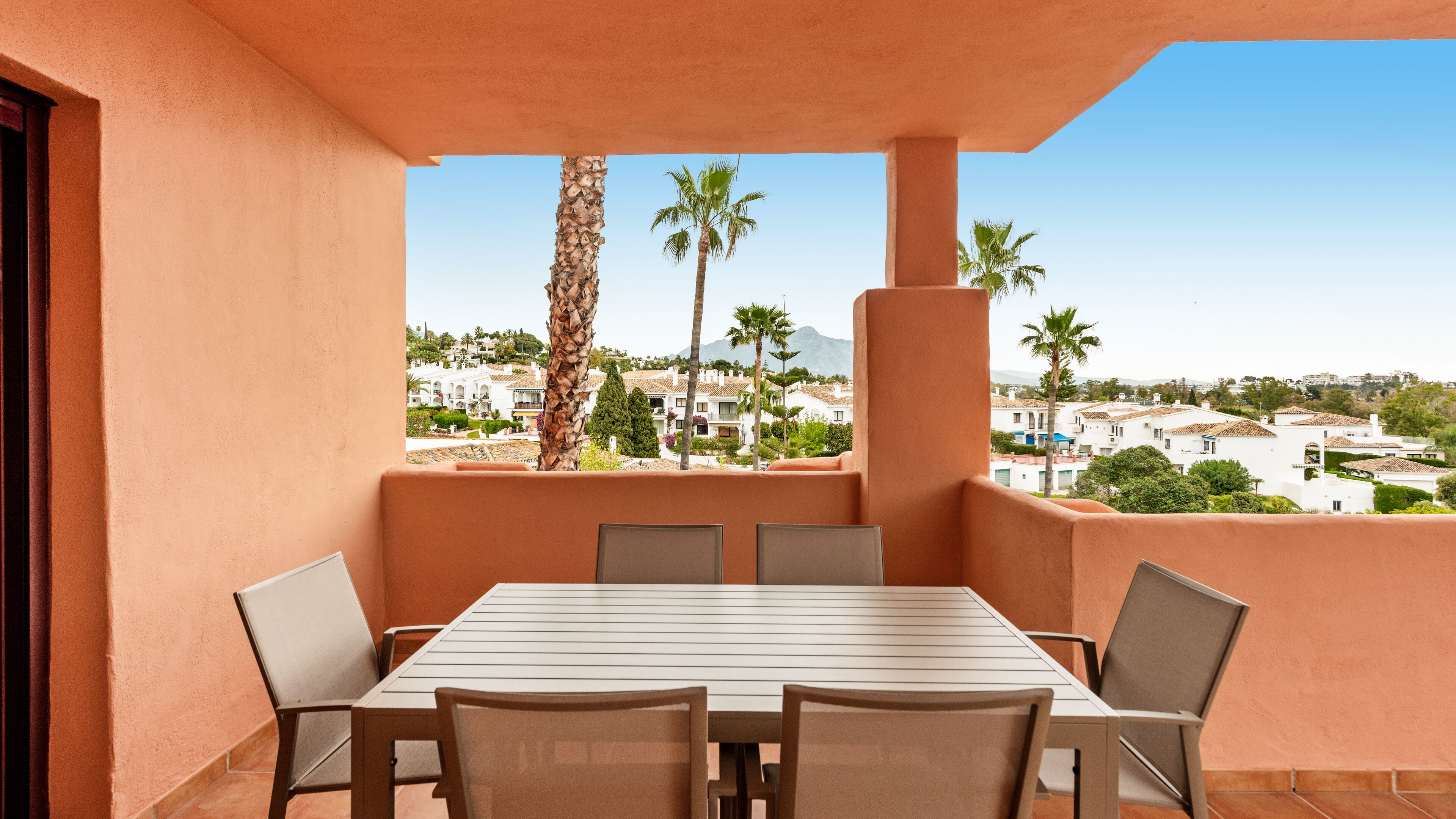 A balcony with a table and chairs, overlooking a residential area with palm trees and houses.