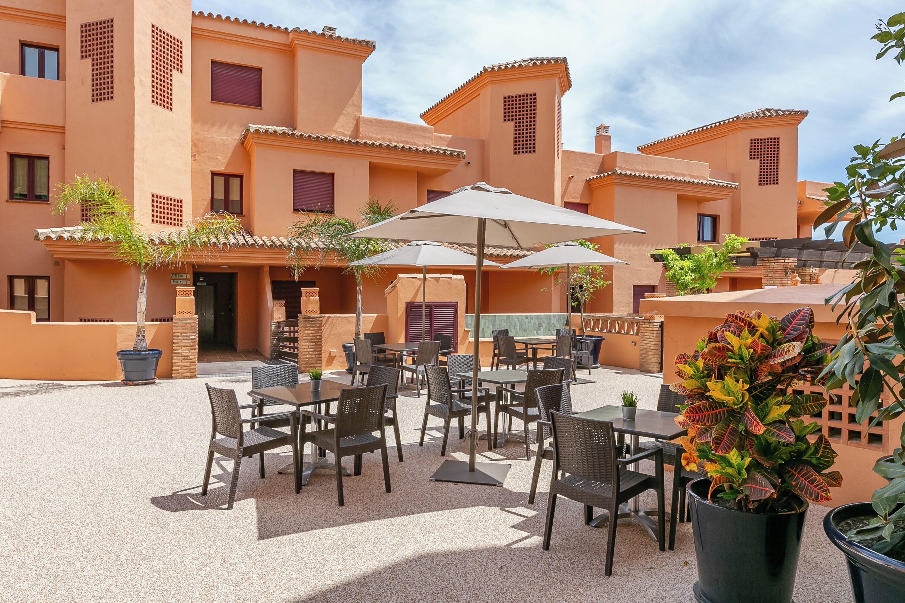 An outdoor patio with tables and chairs, surrounded by buildings with terracotta-colored walls and tiled roofs.