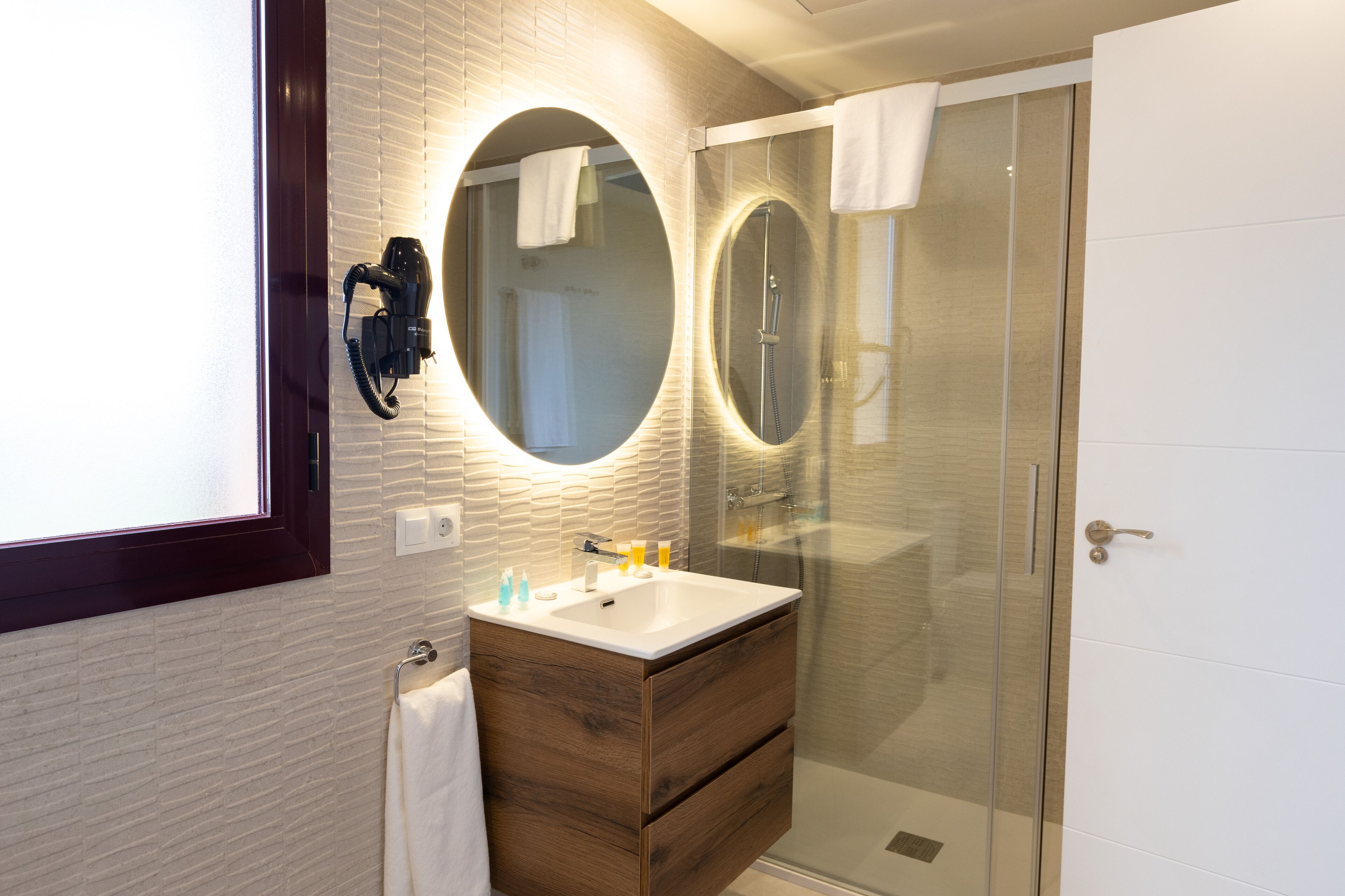 A modern bathroom with a wooden vanity, two round mirrors, a glass shower enclosure, and a hairdryer mounted on the wall.