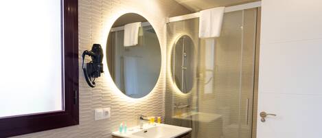 A modern bathroom with a wooden vanity, two round mirrors, a glass shower enclosure, and a hairdryer mounted on the wall.