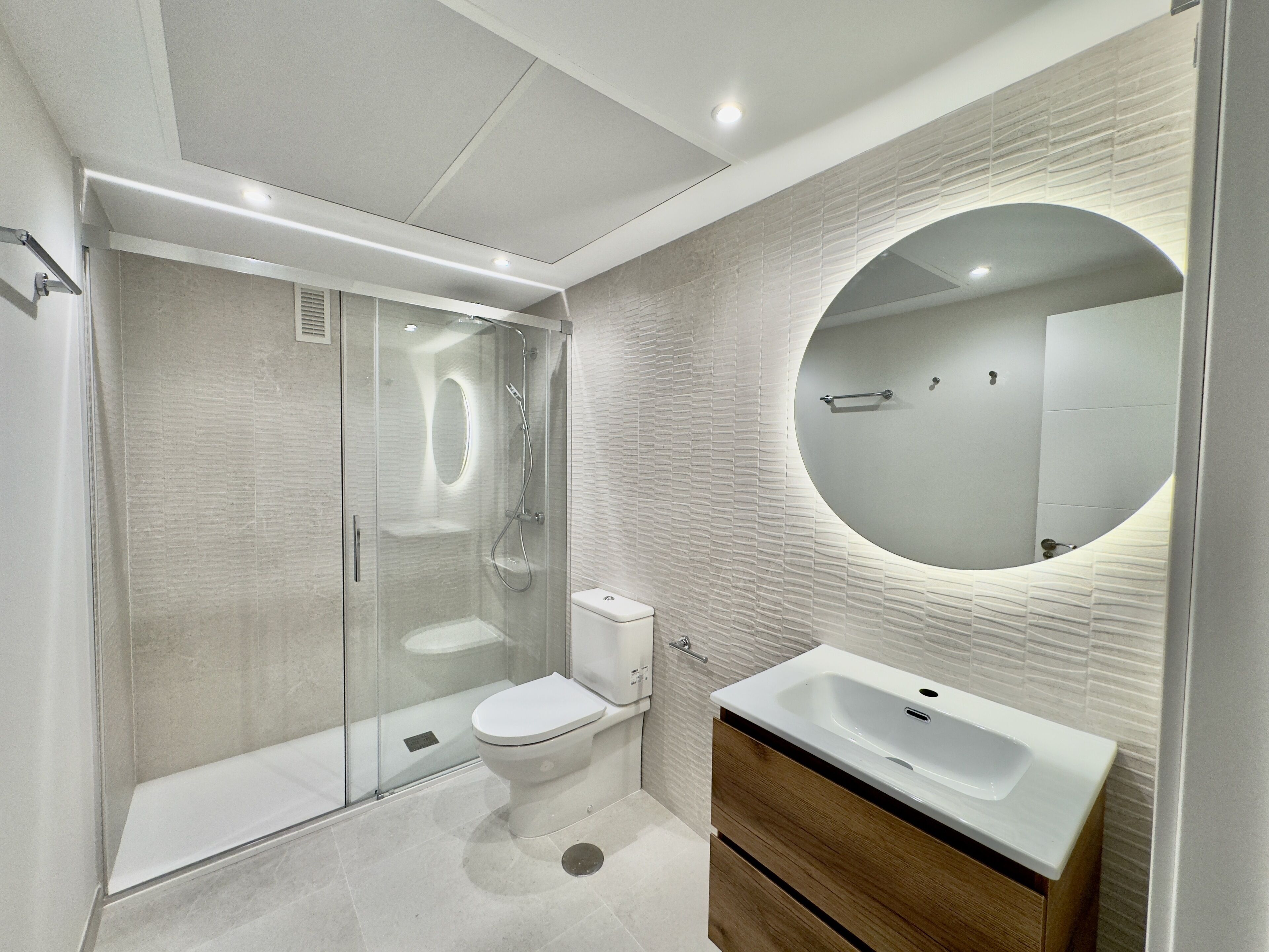 Un bagno moderno con doccia vetrata, un water, un lavandino e uno specchio con cornice rotonda.
