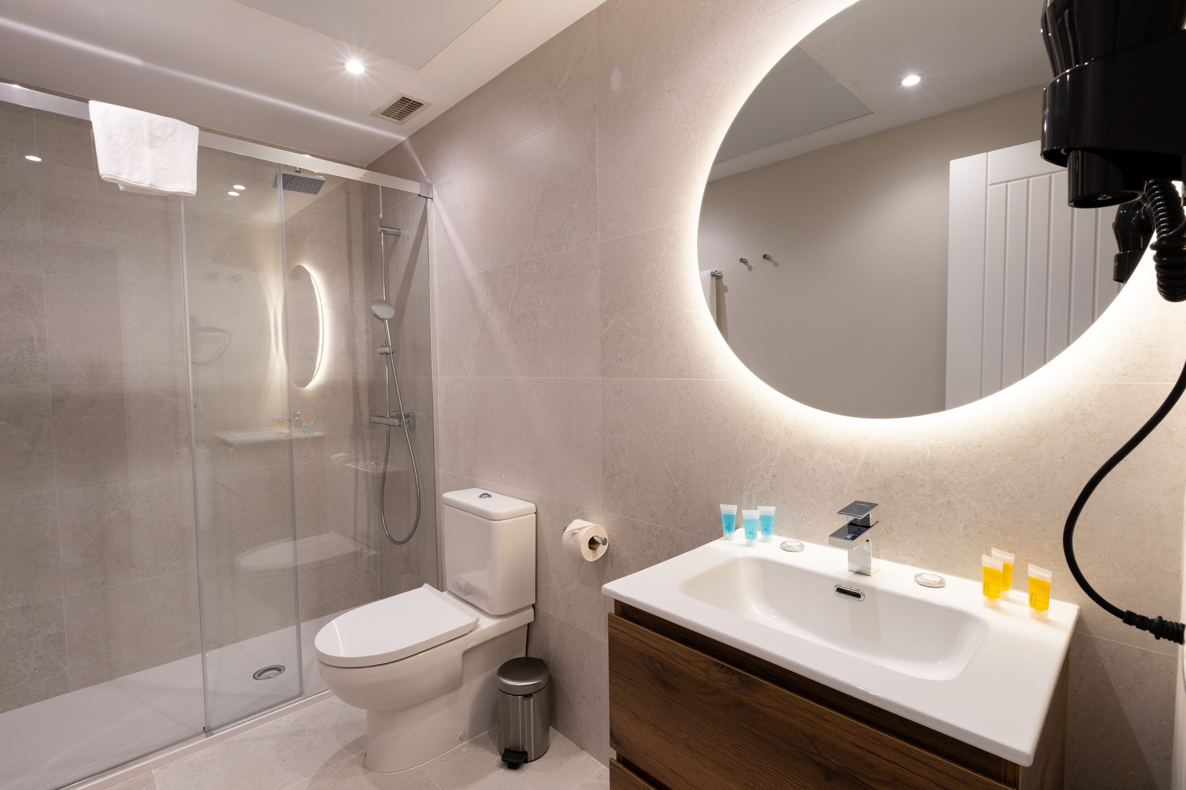 Een moderne badkamer met een douchecabine van glas, een toilet, een wastafel met toiletartikelen en een spiegel met ronde verlichting.