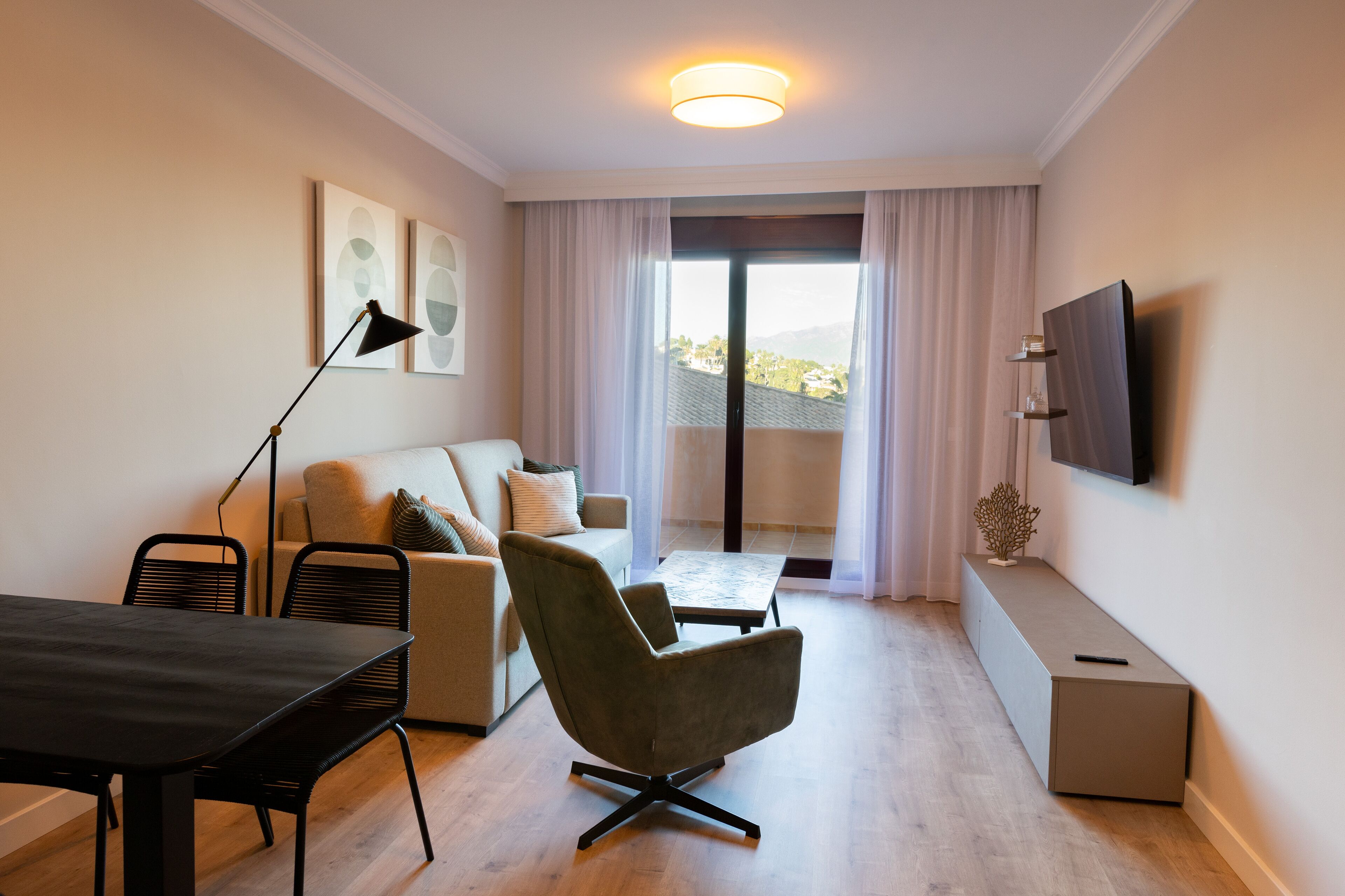 A modern living room with a sofa, armchair, dining table, and a TV mounted on the wall.