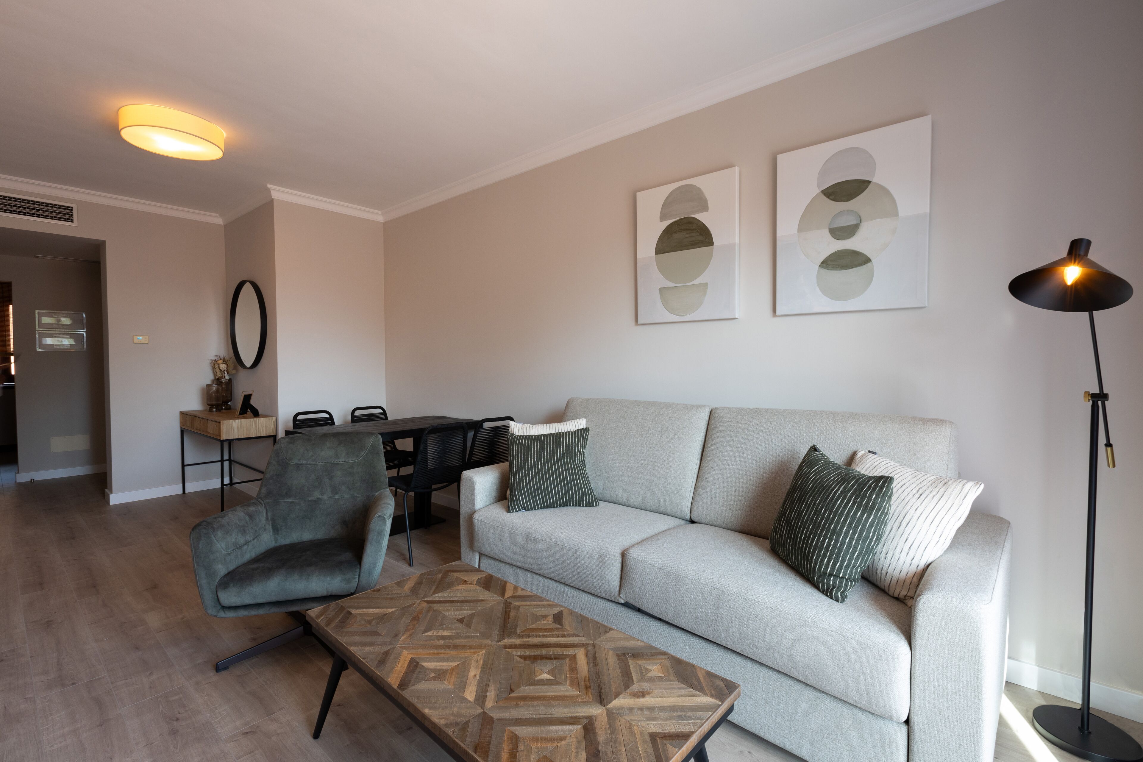 A modern living room with a grey sofa, a patterned coffee table, a grey armchair, and a dining area in the background.