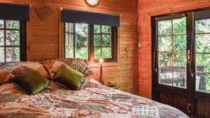 A cozy wooden cabin room with a large bed, a bedside table with a lamp, and a window overlooking greenery.