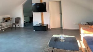 A spacious loft with a high ceiling, a flat-screen TV mounted on a wooden beam, a cozy seating area with a table and cushions, and a view of the sky through a skylight. - roof terrace flat "Alois" (Passau)