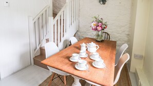 Un comedor con una mesa de madera dispuesta para tomar té, sillas blancas, un jarrón con flores y una escalera con barandillas blancas.