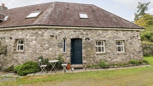 A stone cottage with a dark door, two windows, and a small porch with chairs and a table.