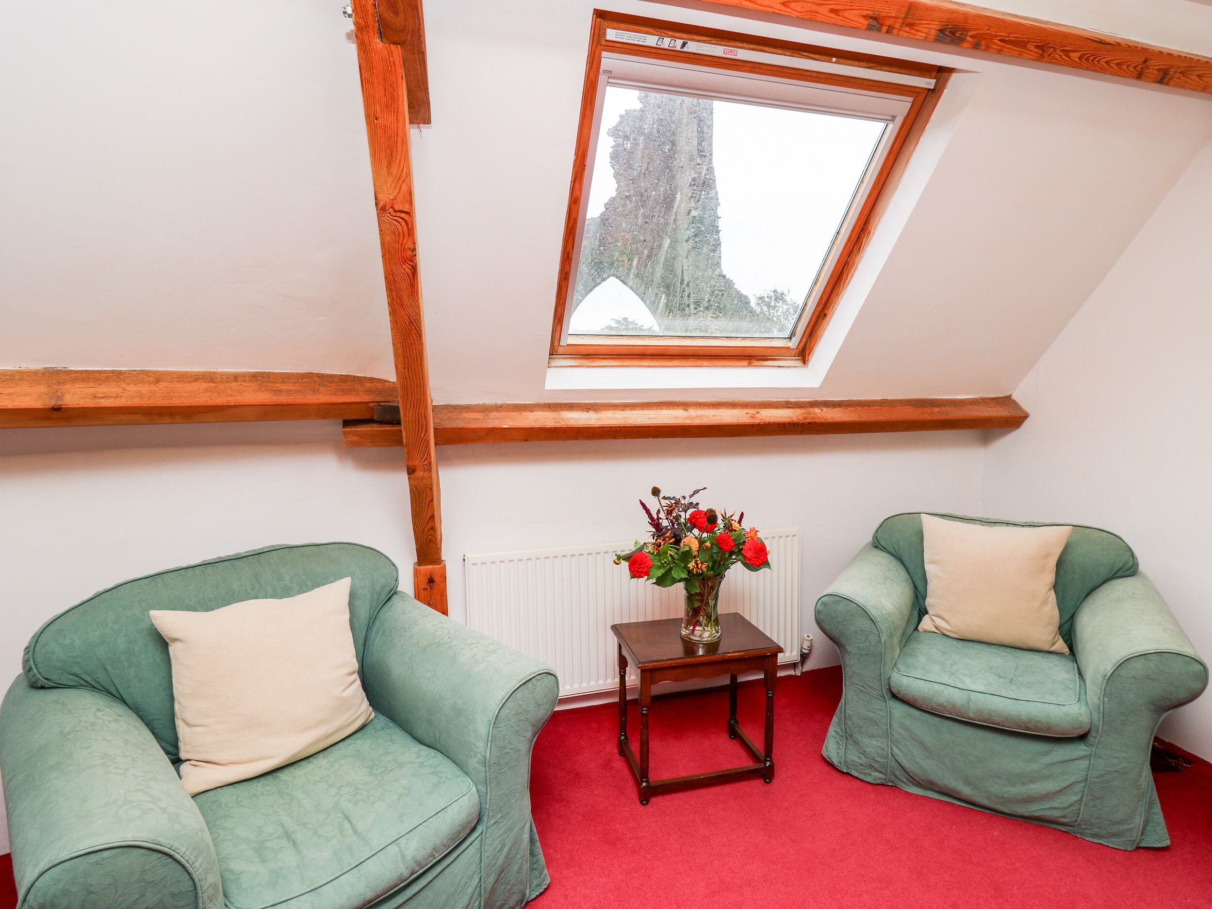 A room with a sloped ceiling, wooden beams, two green armchairs with beige cushions, a small wooden table with a vase of flowers, and a red carpet.