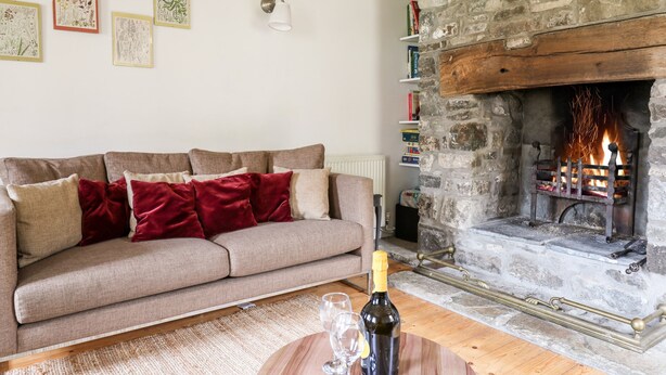 A cozy living room with a stone fireplace, a sofa with red cushions, and framed botanical prints on the wall.