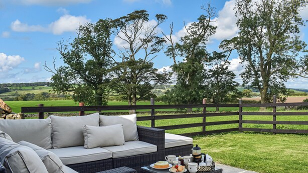 An outdoor seating area with a sofa and wicker furniture, a table with a cake and drinks, and a view of a green field and trees. - 3 bedroom accommodation in Lancaster (Lancaster)