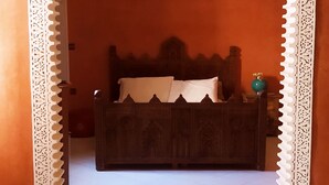 A bedroom with a bed centered under an ornate archway, a nightstand with a lamp, and a wall-mounted light fixture.