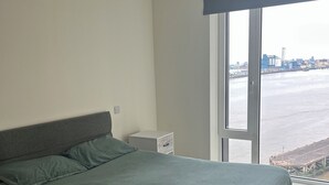 Une chambre à coucher avec un grand lit, une table de chevet, une commode et une vue sur le paysage urbain par la fenêtre.