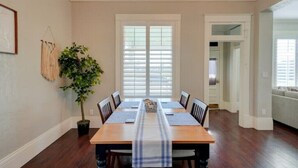 A dining room with a wooden table, six chairs, a striped table runner, a potted plant, and a window with blinds.