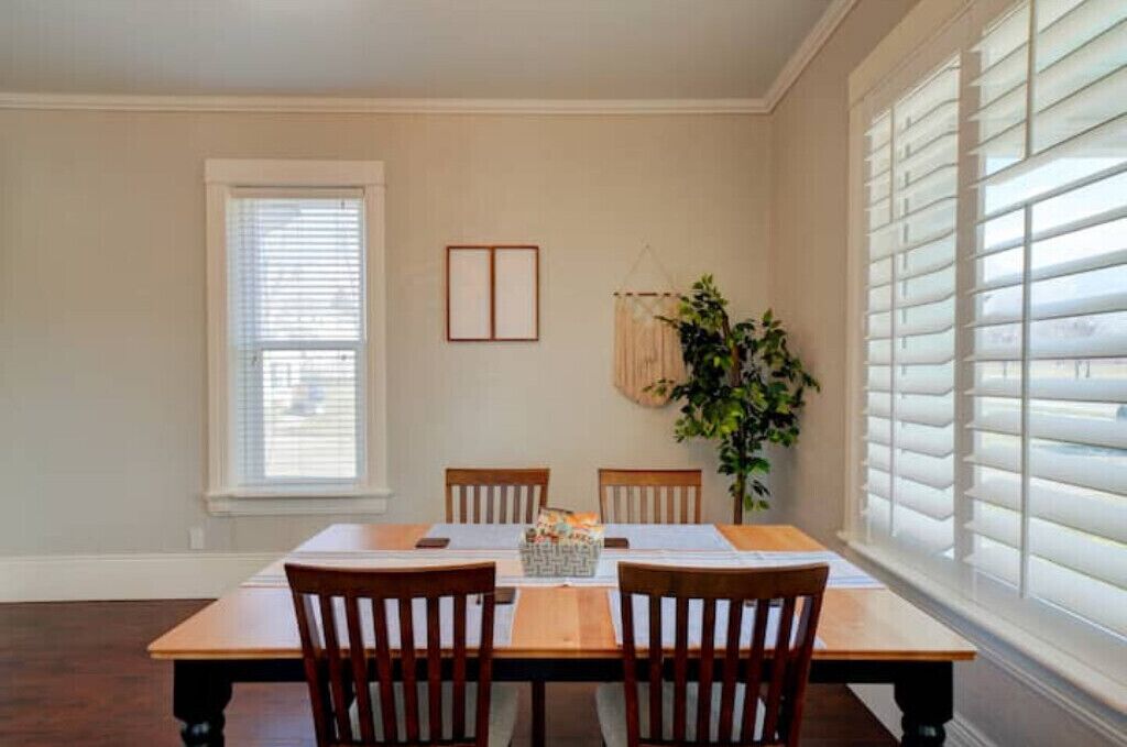 A dining room with a wooden table and chairs, a window with blinds, and a hanging plant.