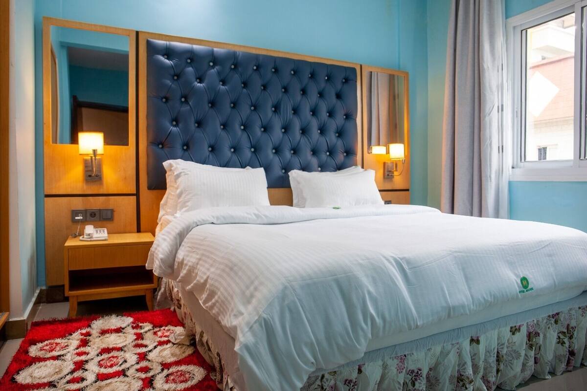 A hotel room with a large bed, a tufted headboard, bedside lamps, a wooden nightstand, and a red and white patterned rug.
