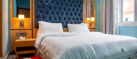 A hotel room with a large bed, a tufted headboard, bedside lamps, a wooden nightstand, and a red and white patterned rug.