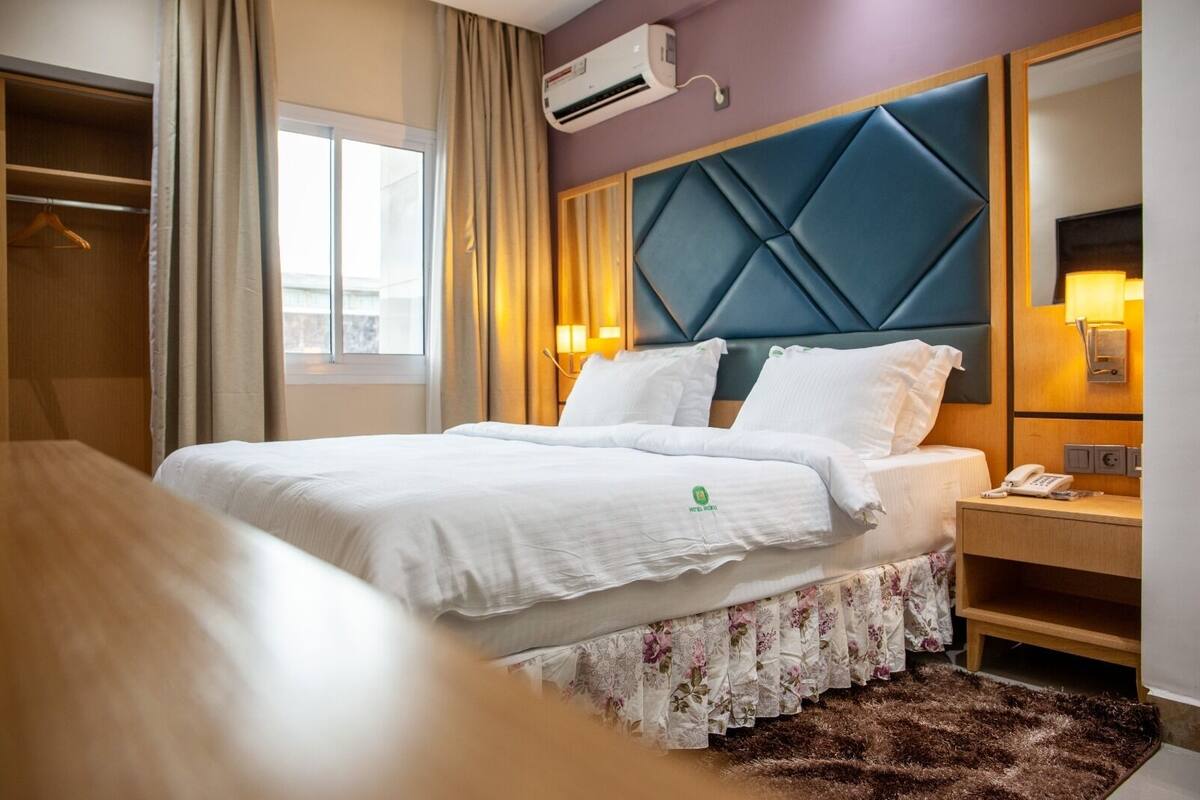 A hotel room with a bed, a wooden nightstand, a mirror, and an air conditioning unit.