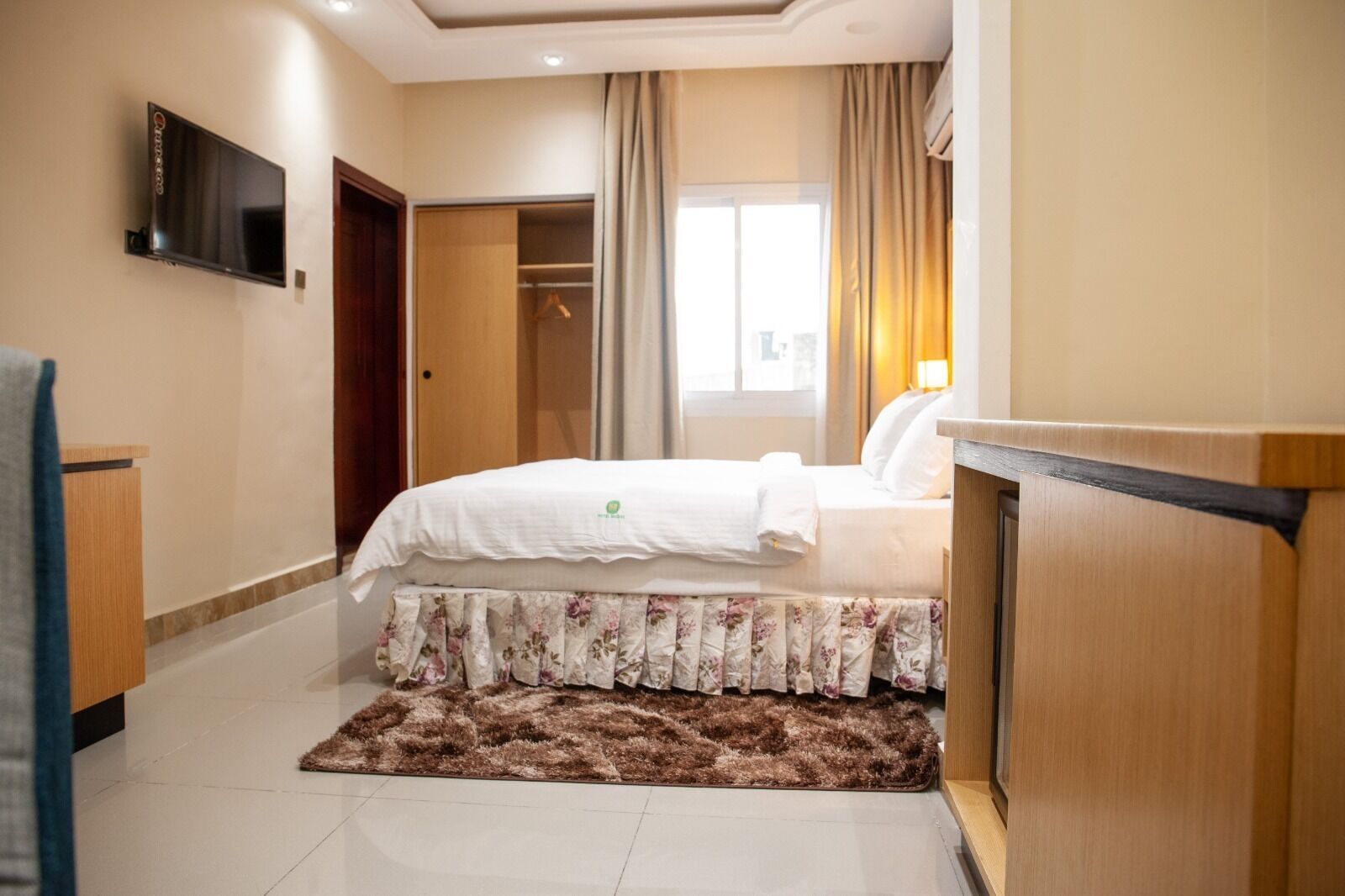 Habitación de hotel con cama, televisor montado en la pared, escritorio de madera, armario y ventana con cortinas.