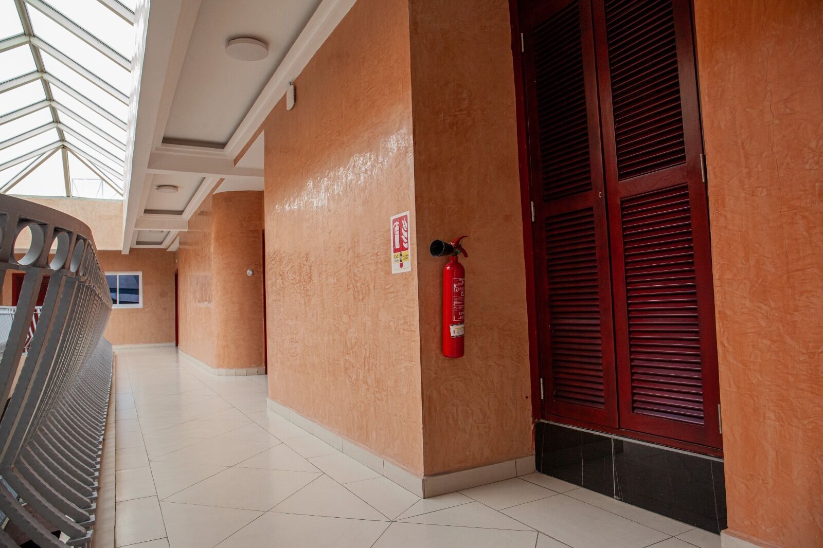 A corridor with a fire extinguisher mounted on the wall, a fire alarm sign, and a door with red louvered shutters.