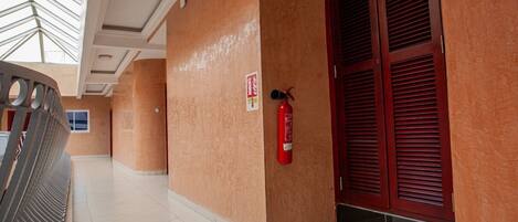 A corridor with a fire extinguisher mounted on the wall, a fire alarm sign, and a door with red louvered shutters.