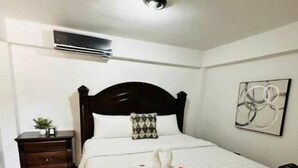 Una cama bien hecha con un cabecero oscuro, una mesita de noche pequeña con una lámpara y un aire acondicionado de pared.