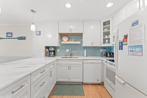 A modern kitchen with white cabinets, a marble countertop, a built-in dishwasher, and a coffee maker. - Beachmaster 104-2BR Oceanfront Condo (North Myrtle Beach)