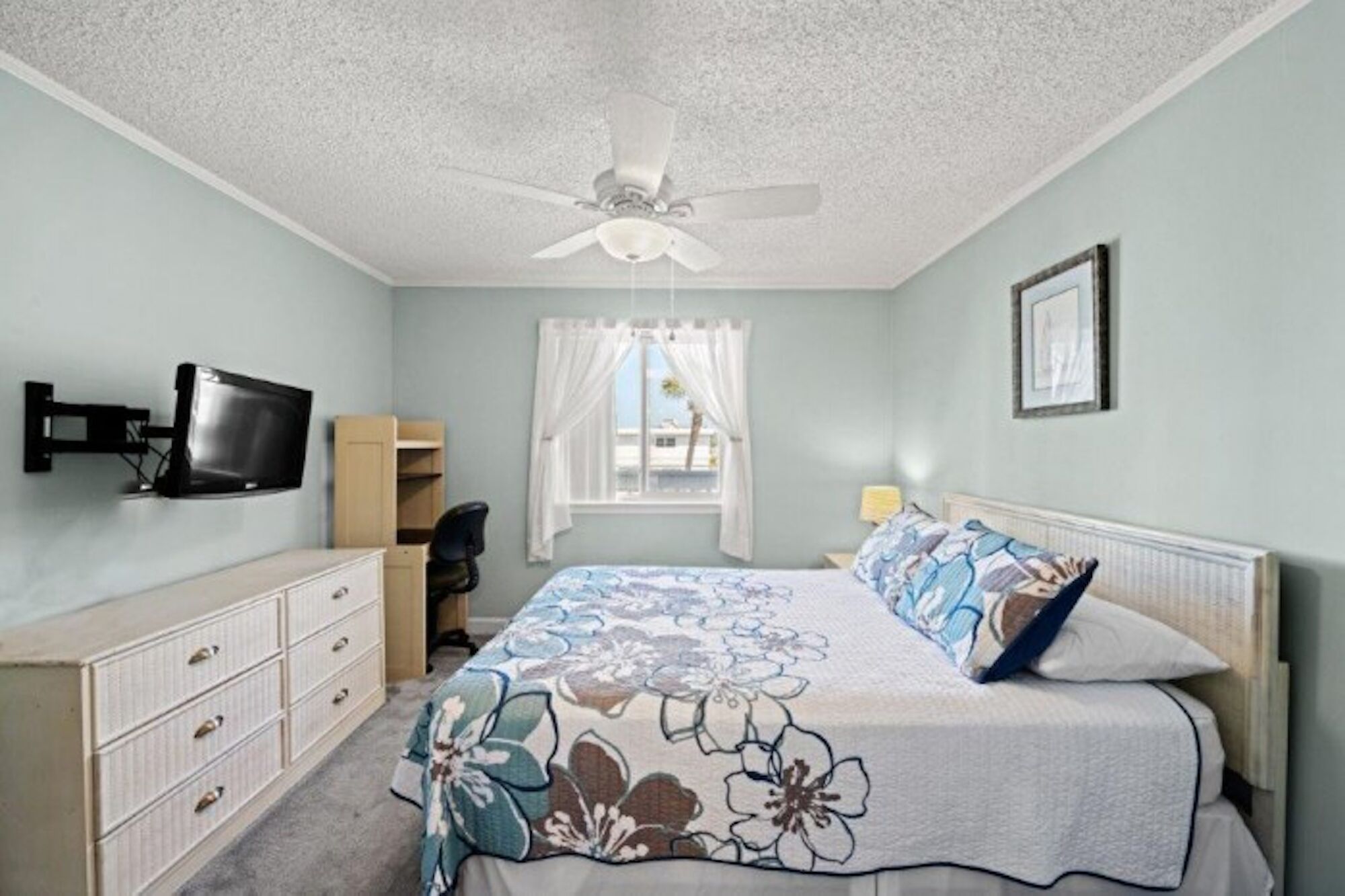 A bedroom with a bed, a desk, a chair, a television, a window with curtains, and a ceiling fan.