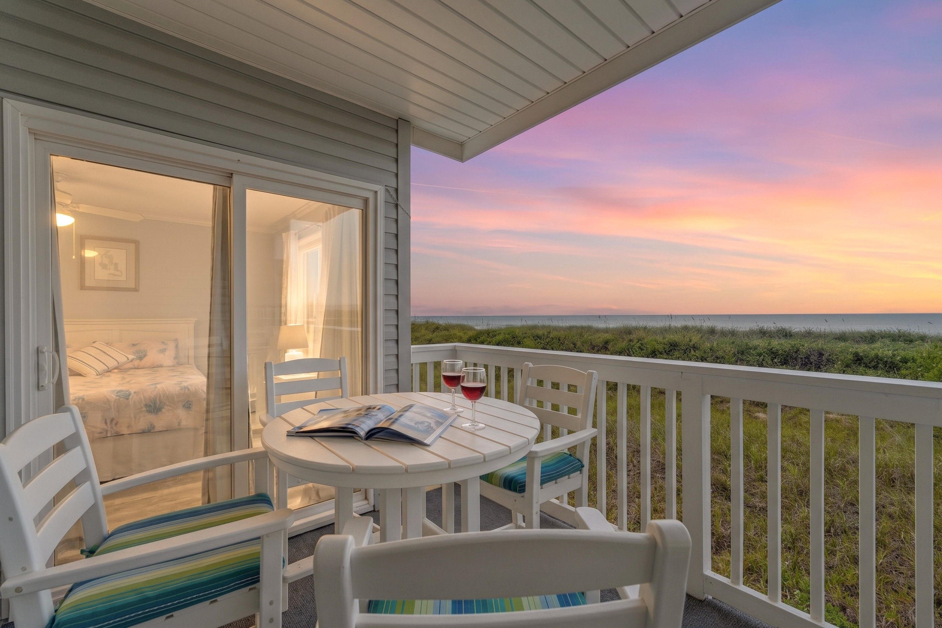 A balcony with a table, chairs, and a book, overlooking a grassy area and a sunset.