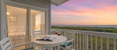 A balcony with a table, chairs, and a book, overlooking a grassy area and a sunset.