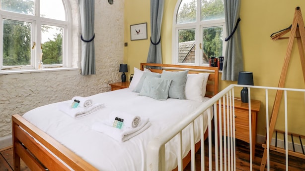 A bedroom with a wooden bed, two windows with curtains, a bedside table with a lamp, and a wooden ladder against the wall. - Hapton Folly (Waterlooville)