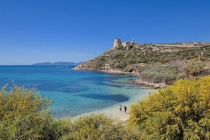 Un paysage côtier avec une mer bleue et limpide, une colline rocheuse surmontée d’une tour et une végétation clairsemée.