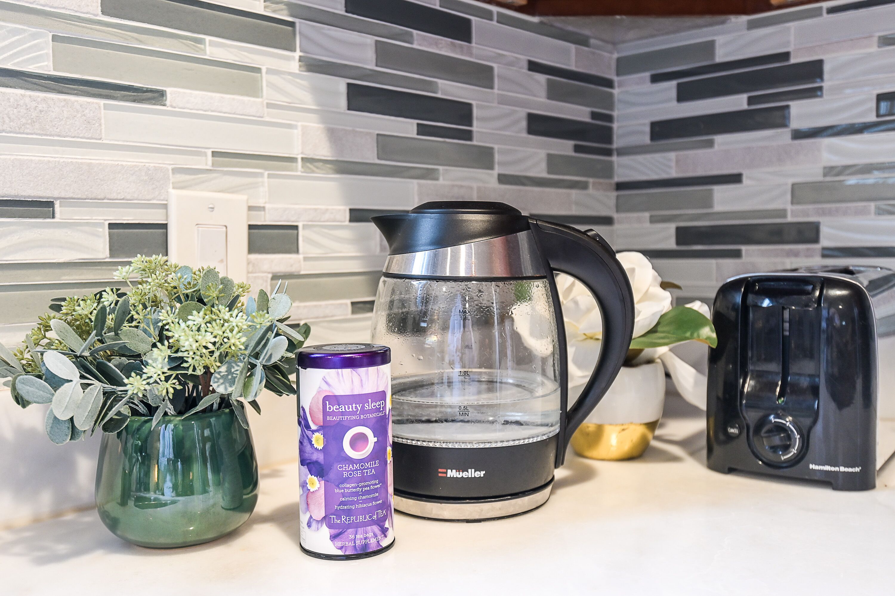A kitchen counter with a green vase, a can of tea, a black coffee maker, and a toaster.