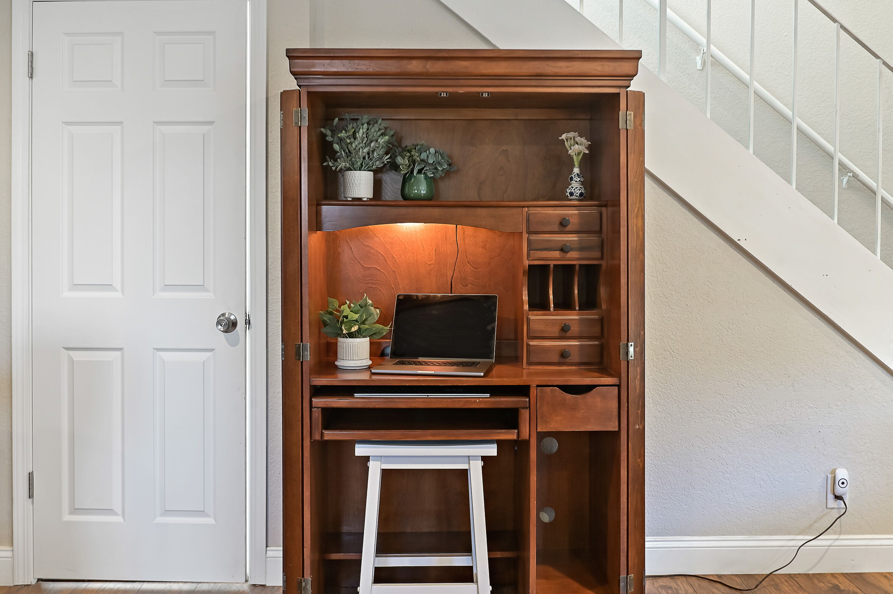 A compact wooden desk with a laptop, a small plant, and a vase with flowers.