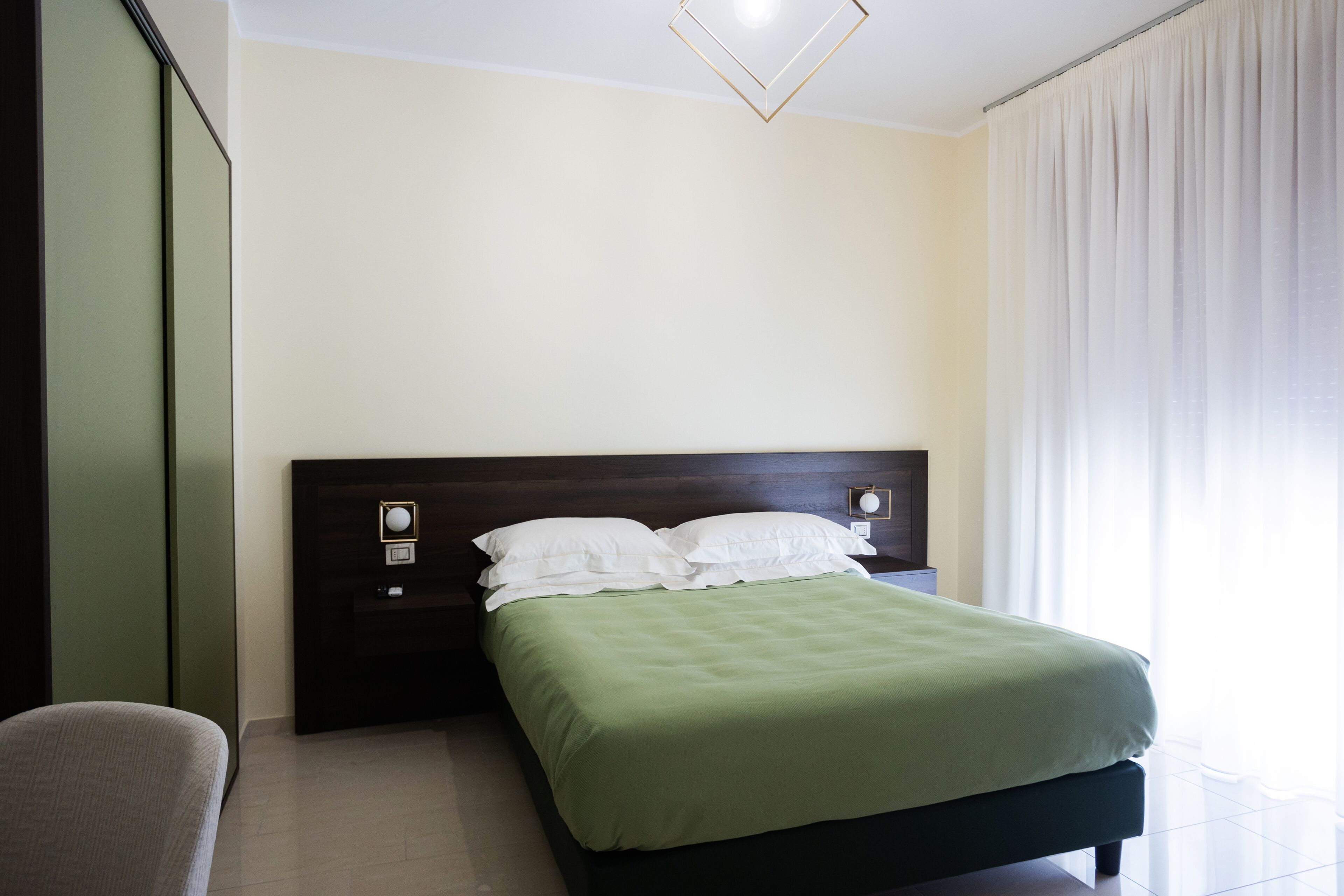 A neatly made bed with a green bedspread and white pillows in a modern room with a dark headboard, a bedside table with a lamp, and a large window with curtains.