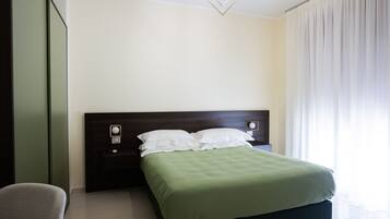 A neatly made bed with a green bedspread and white pillows in a modern room with a dark headboard, a bedside table with a lamp, and a large window with curtains.