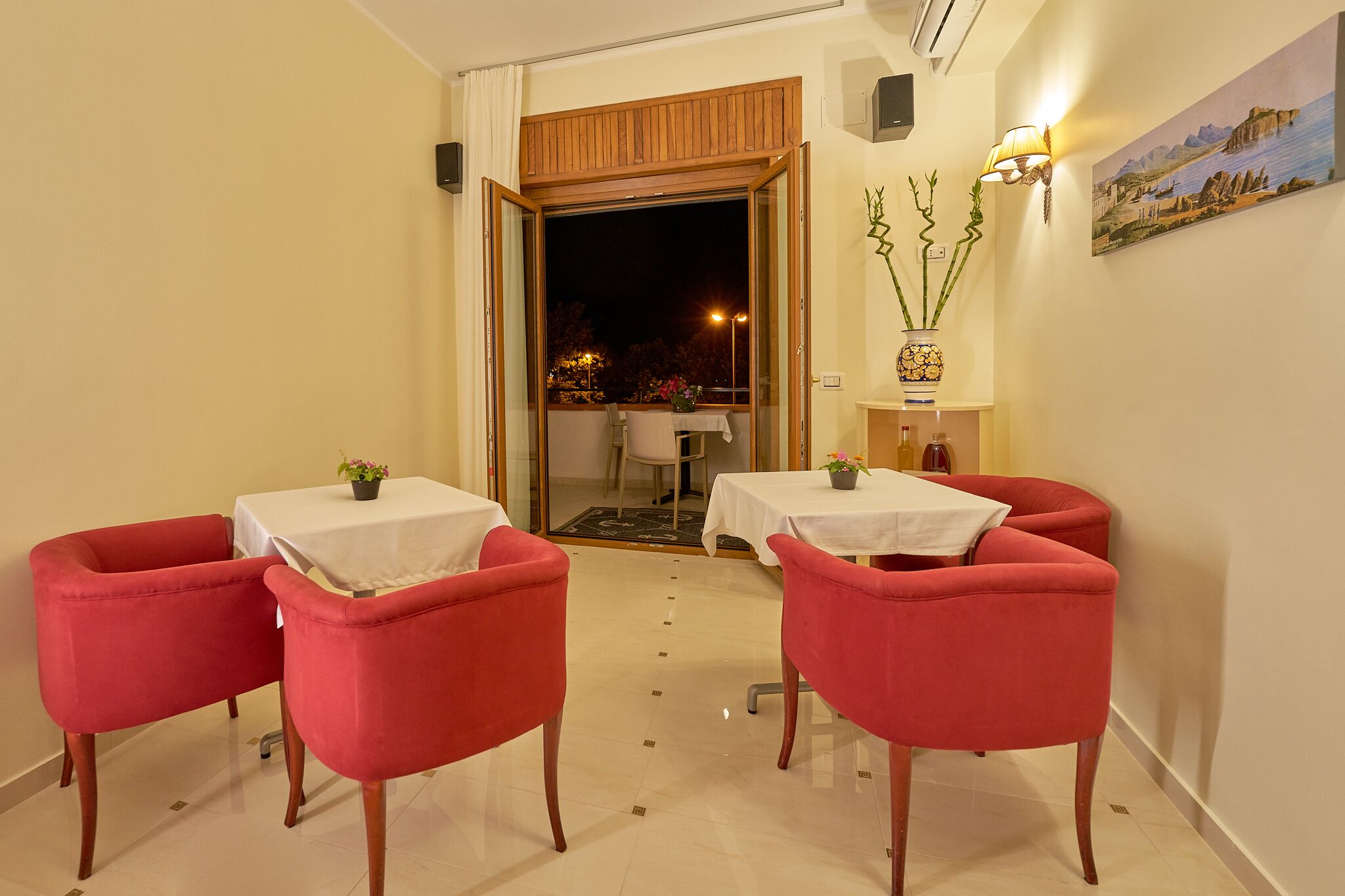 Un coin repas avec deux chaises et une table rouges, une porte en bois donnant sur l’extérieur, une plante en pot et un tableau accroché au mur.