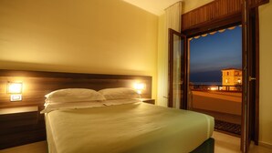 A hotel room with a bed, nightstands, and a view of the sea at dusk.