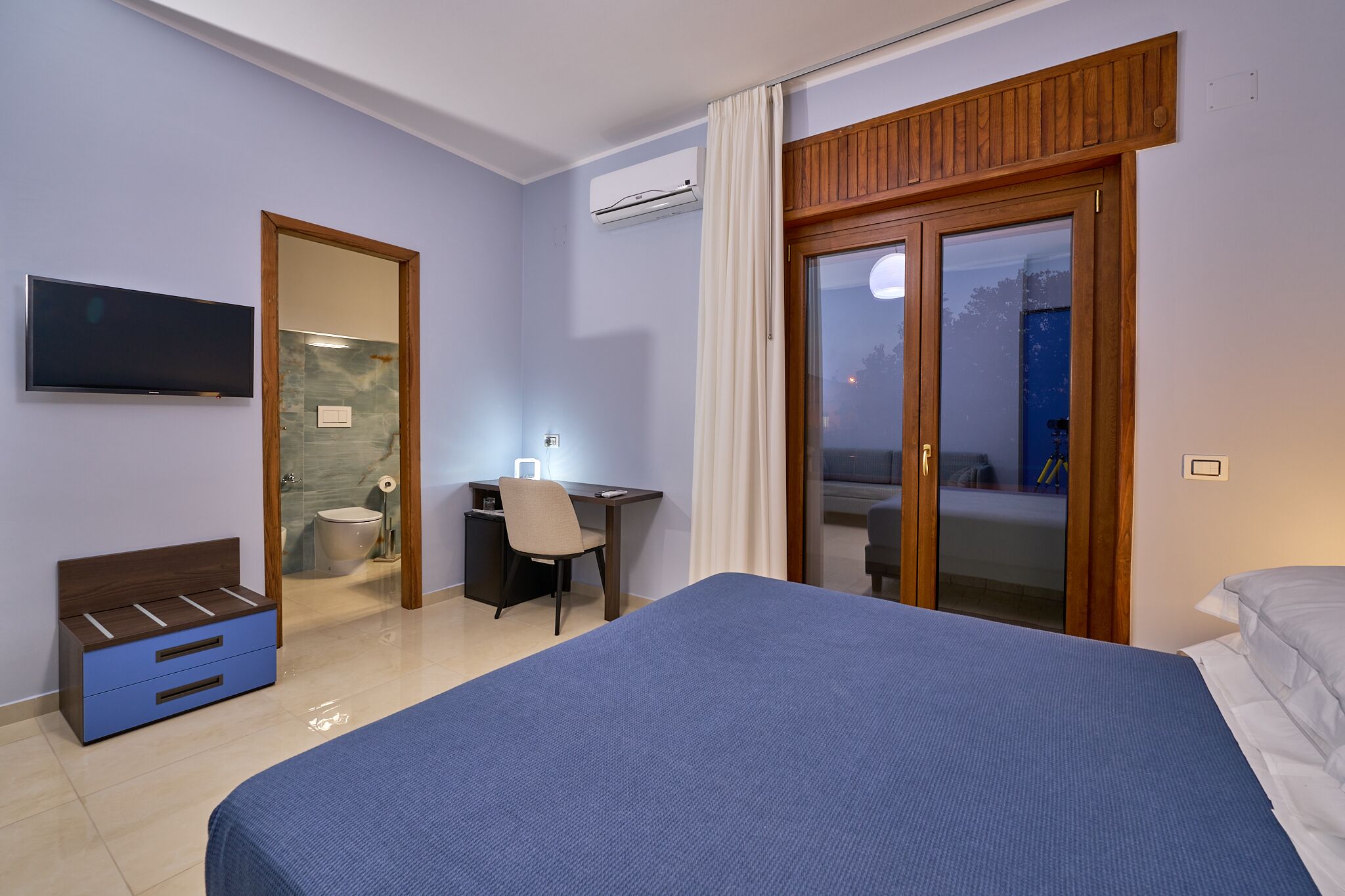 A hotel room with a bed, a desk, a chair, a television, and a sliding glass door leading to an outdoor area.