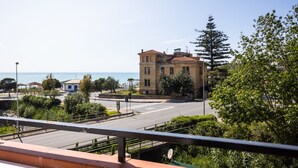 A coastal building with a view of the sea, surrounded by greenery and a clear sky.