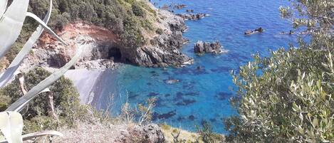 A coastal landscape with a rocky beach, clear blue waters, and a cave entrance.