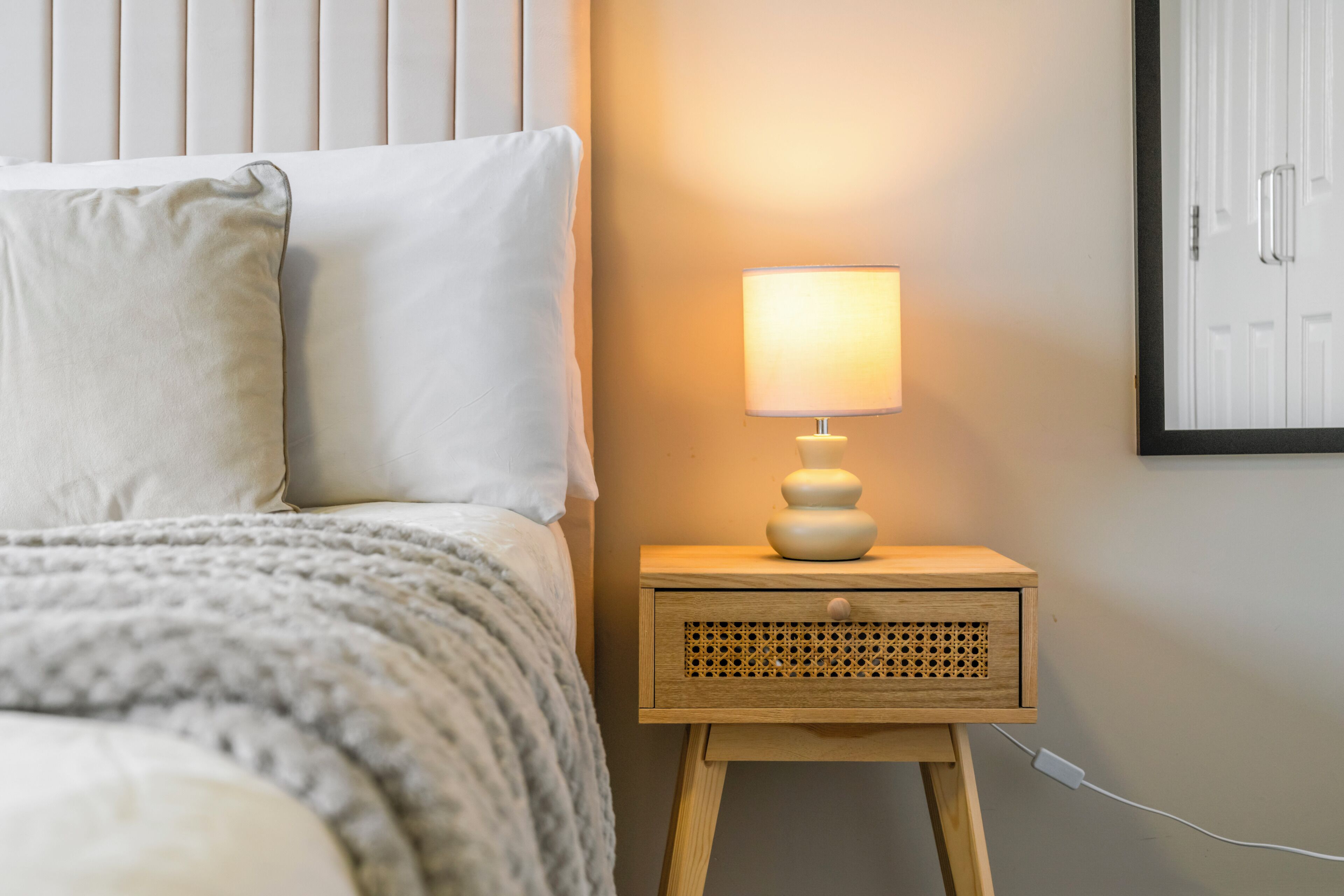 Uma cama bem arrumada com travesseiro branco e manta cinza, uma mesa de cabeceira de madeira com abajur, e uma porta fixada na parede.