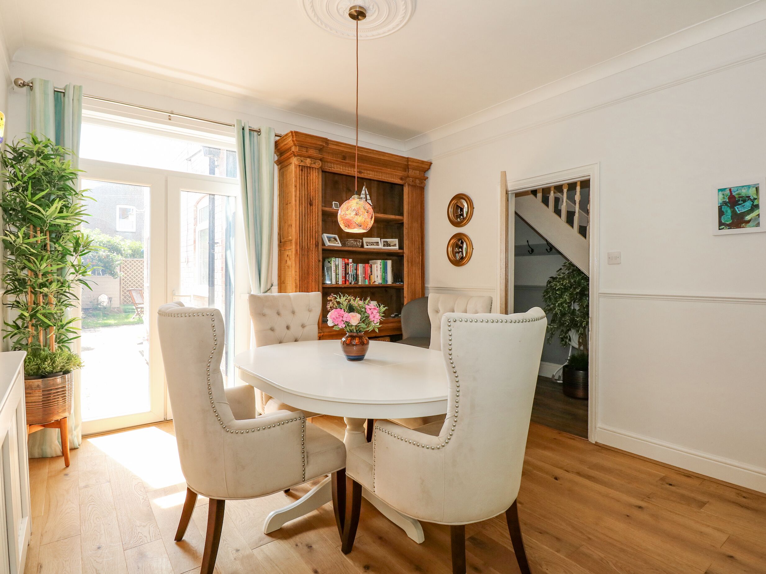 Comedor con mesa redonda, cuatro sillas tapizadas, lámpara colgante y una estantería con libros y objetos decorativos.