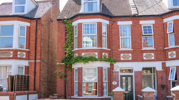 A red brick terraced house with bay windows, a front door, and a small garden area.