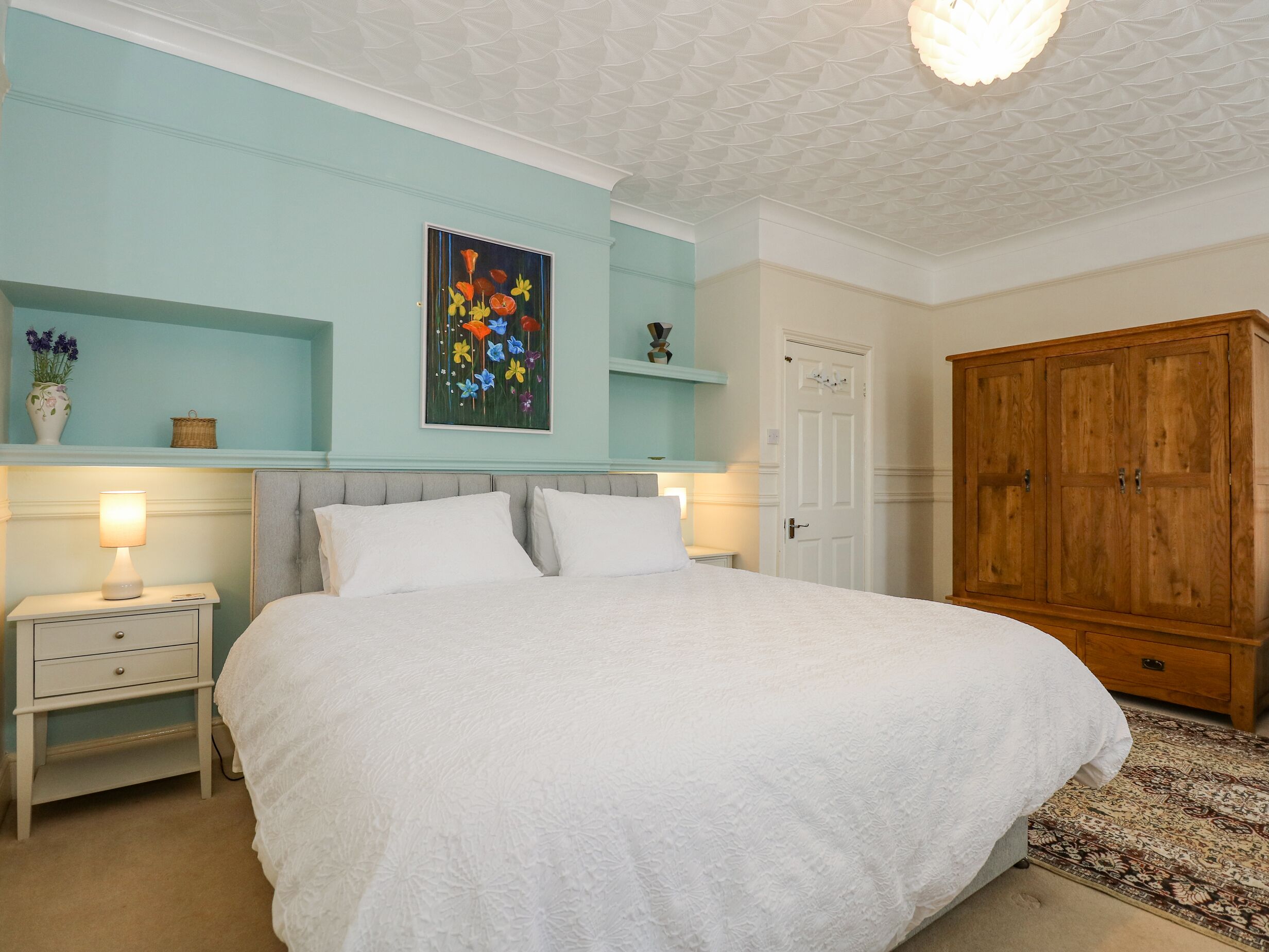 A bedroom with a large bed, a nightstand with a lamp, a wardrobe, and a wall-mounted shelf with decorative items.