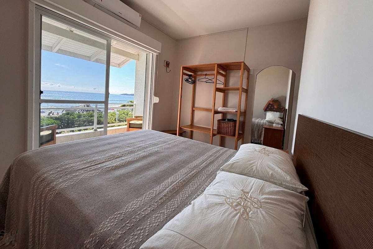 A bedroom with a bed, a wooden wardrobe, a mirror, and a balcony with a view of the sea.